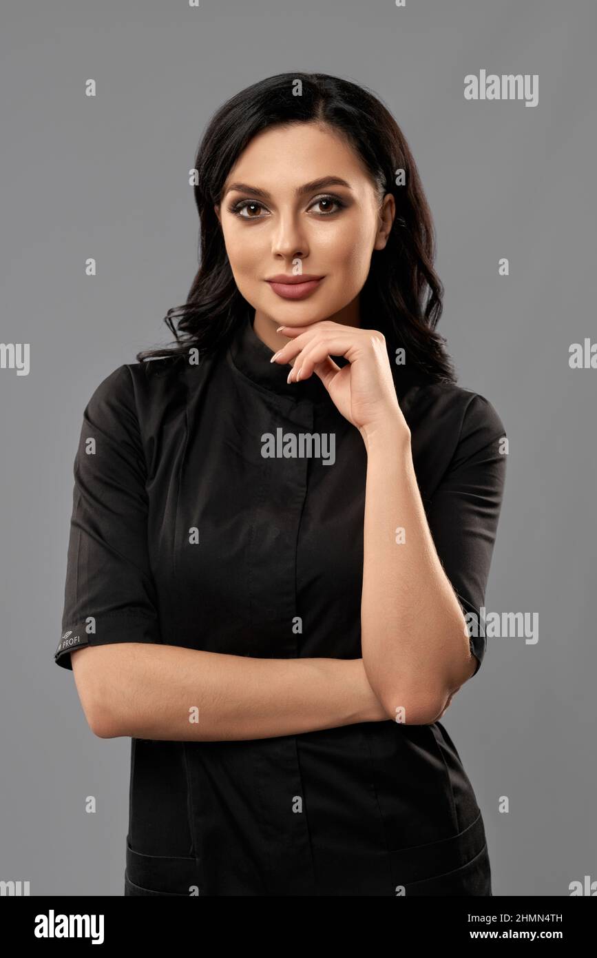 Confident dark haired woman wearing black medical uniform standing in studio with grey background. Competent dermatologist indoors. Beauty and care concept. Stock Photo