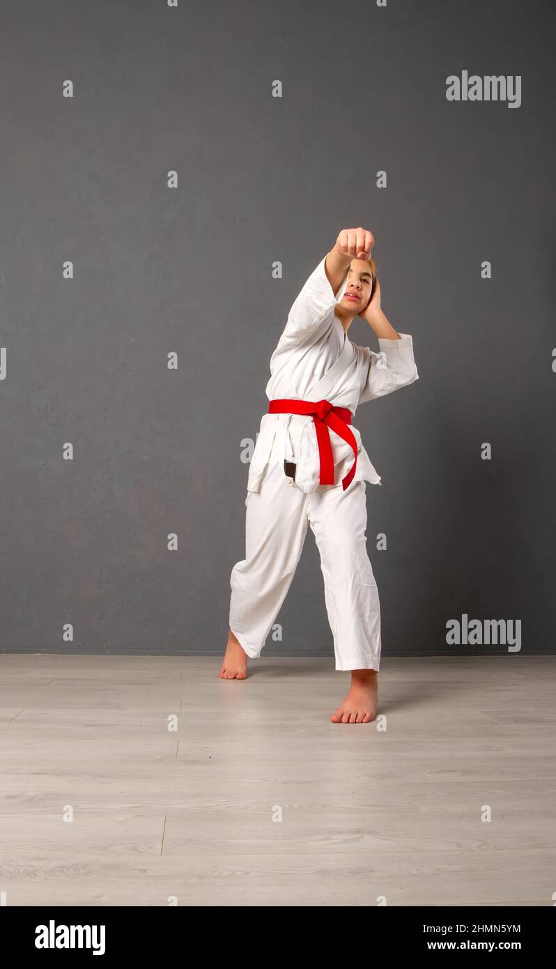 A young girl karateka in a white kimono and a red belt trains and performs a set of exercises against a gray wall Stock Photo