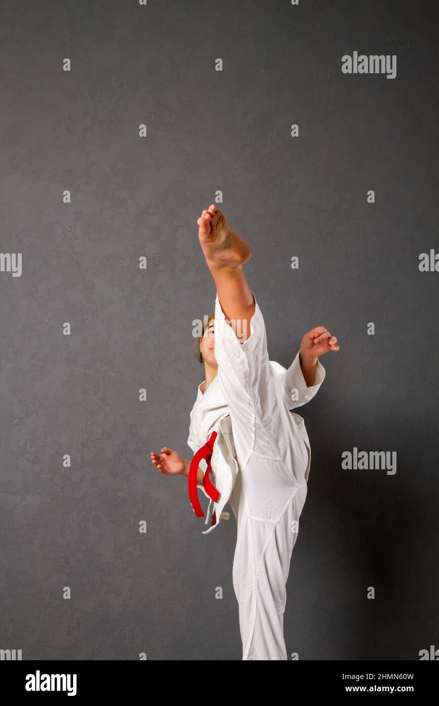 A young girl karateka in a white kimono and a red belt trains and performs a set of exercises against a gray wall Stock Photo