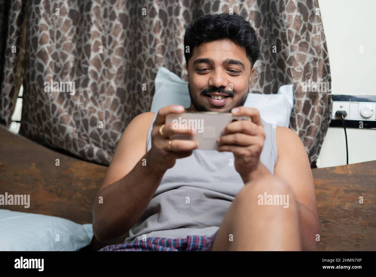 happy young man playing video game on mobile phone at bedroom - concept of entertainment, smartphone addiction and killing time Stock Photo