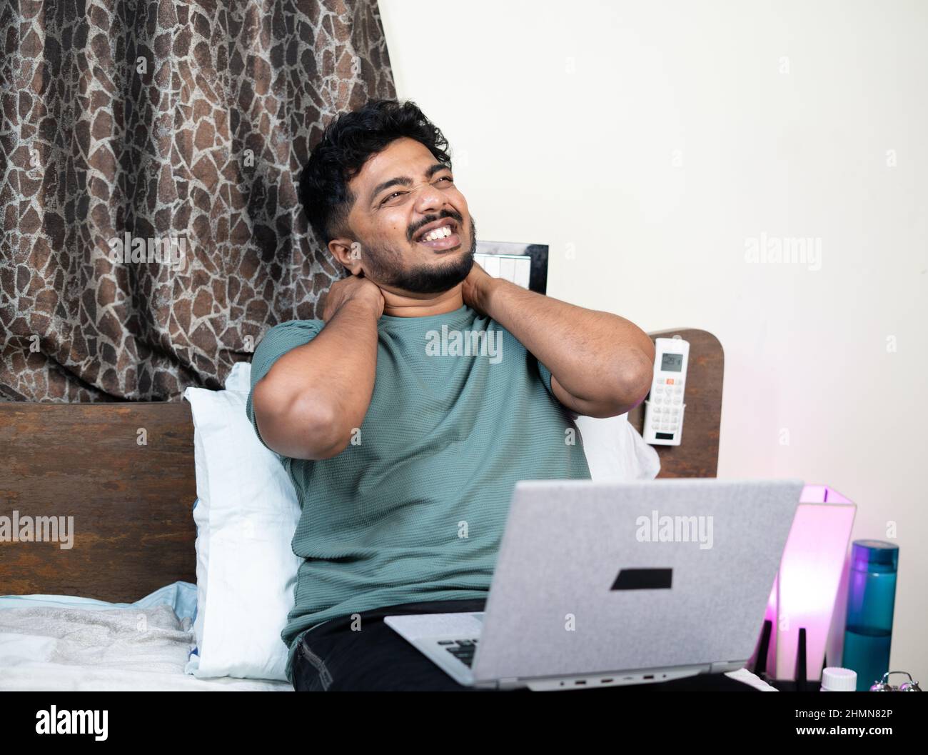 young man suffering from neck and back pain while using laptop on bed during work from home - concept of improper working posture, overworked and Stock Photo