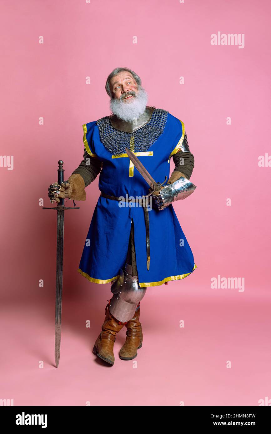 Portrait of senior grey bearded man, brave medieval warrior or knight in  armored clothes with sword isolated on pink background. Comparison of eras  Stock Photo
