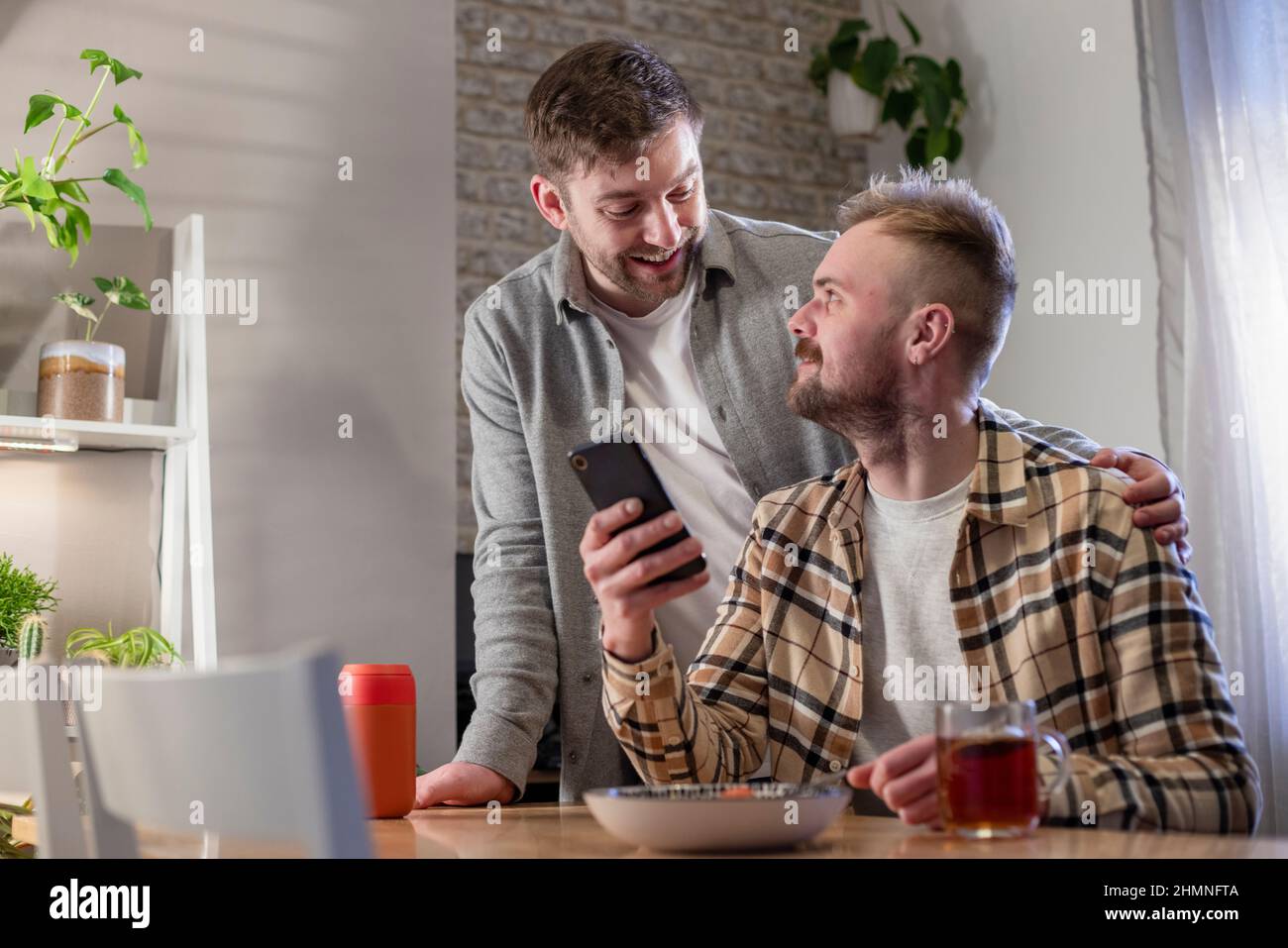 An LGBTQI same sex male couple sitting at a table in their living room relaxing, one man is using his smart phone and he has a plate of pasta. Stock Photo