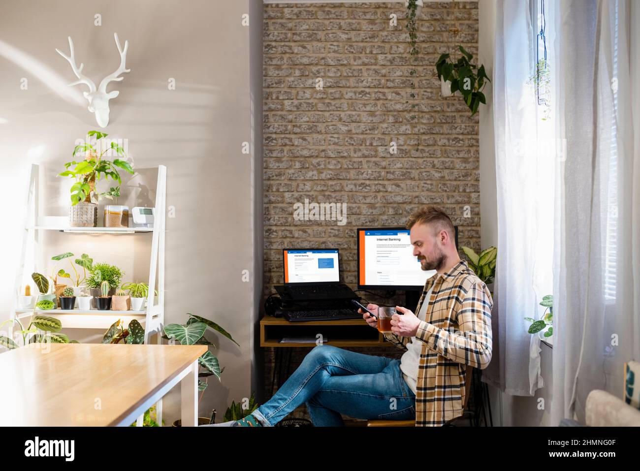 A shot of a home office workspace desk with two monitors, internet banking is displayed on the screens. A man is sitting using his phone by his desk. Stock Photo