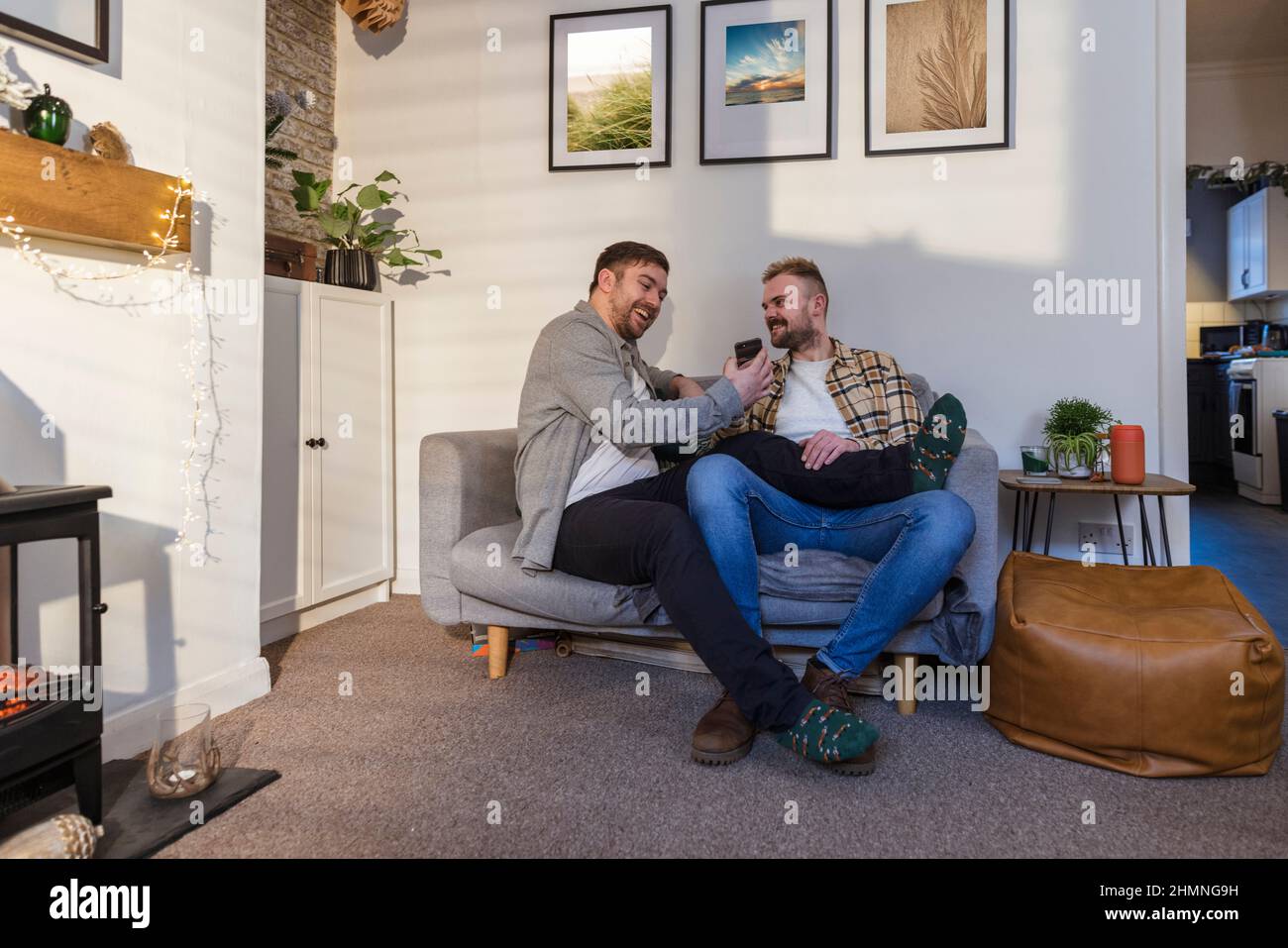 A front-view shot of an LGBTQI same sex male couple sitting in their living room relaxing, one man is showing his partner his smart phone. Stock Photo