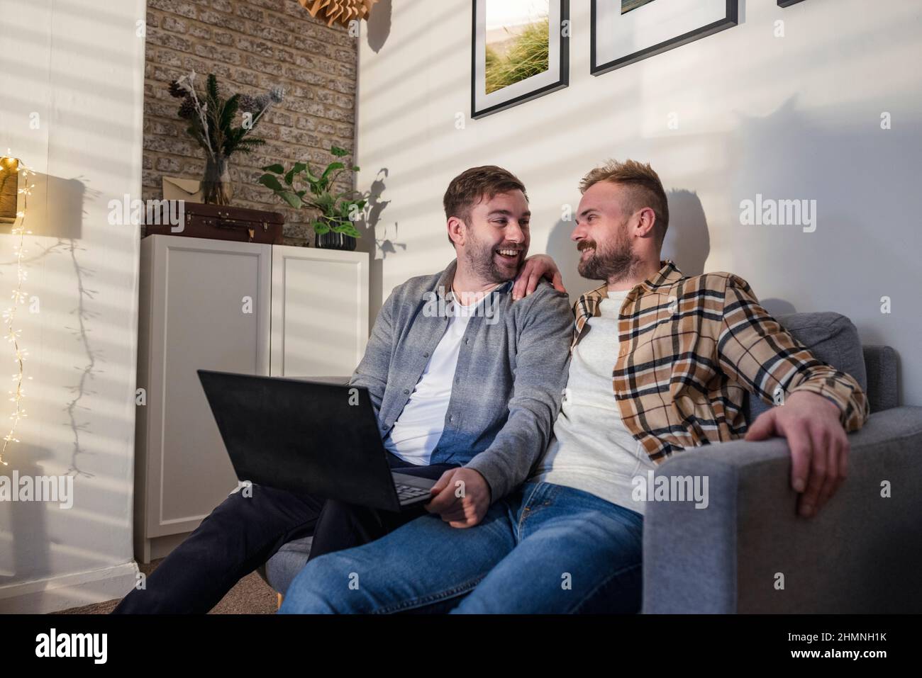 An LGBTQI same sex male couple sitting on their sofa in their living room relaxing, they are using a laptop and smiling to one another. Stock Photo