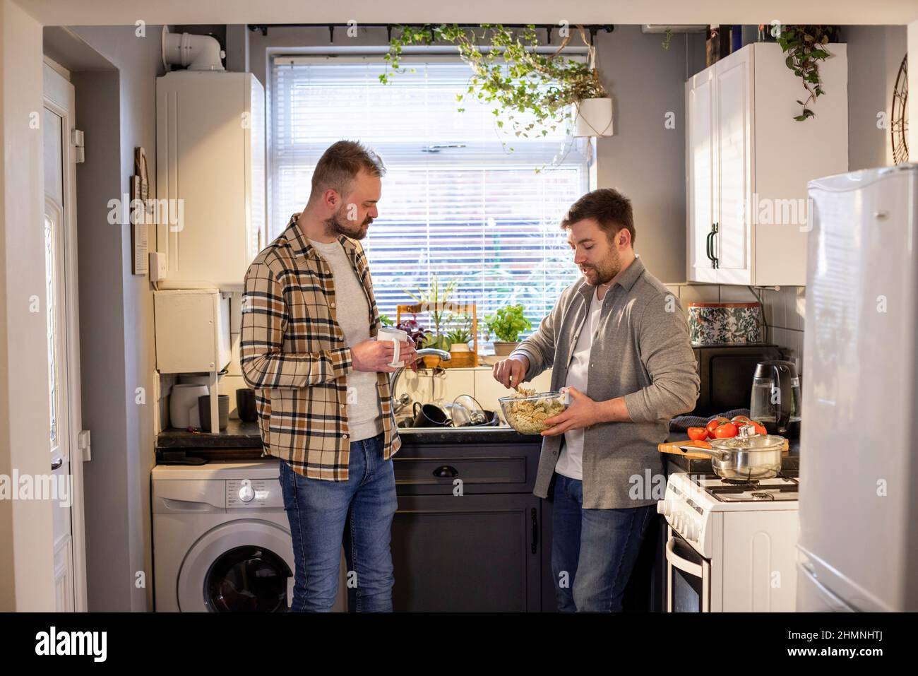 A side-view shot of an LGBTQI same sex male couple standing in their kitchen together, one man is making pasta while the other man talks to him. Stock Photo