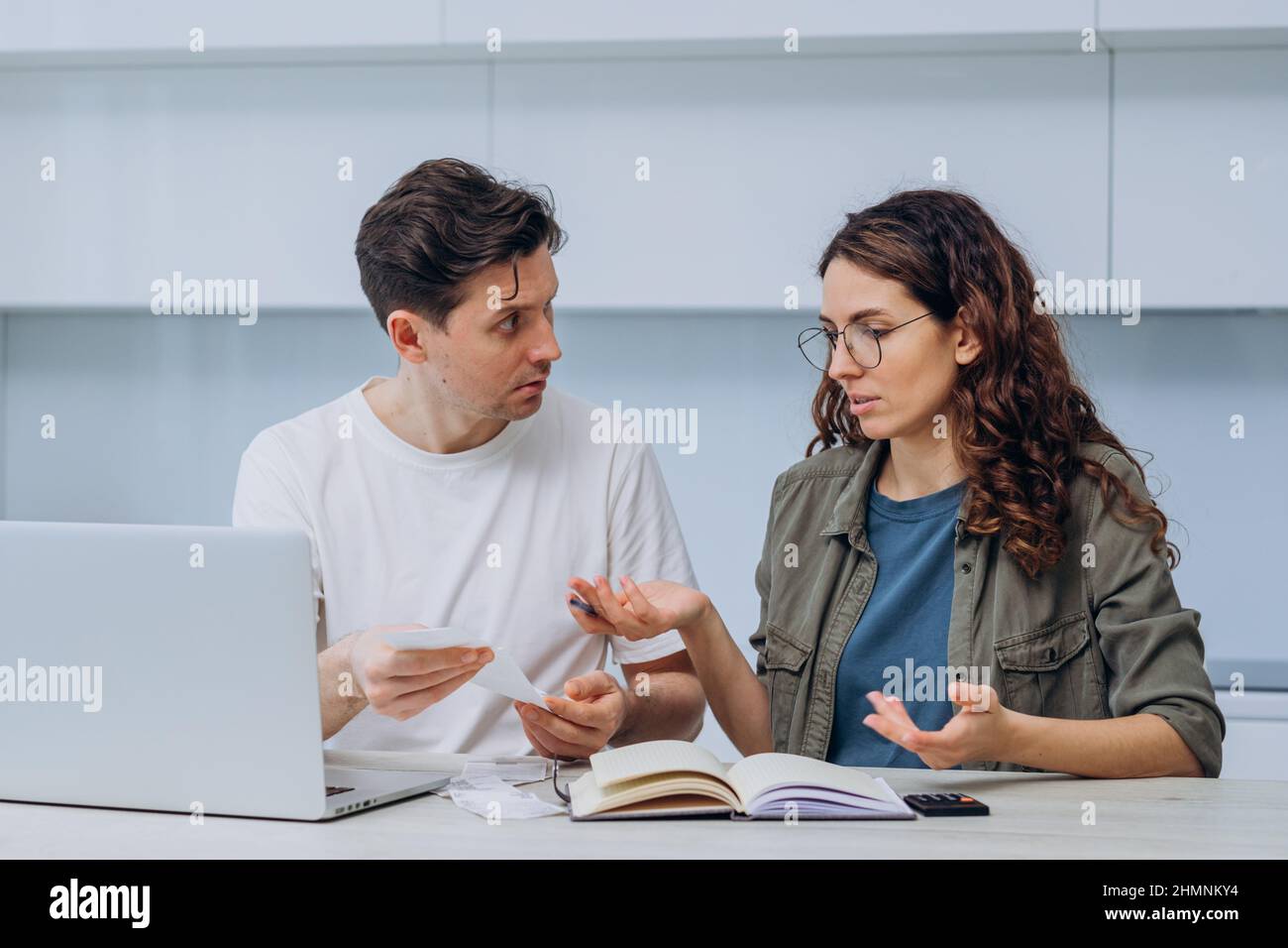 Husband and wife calculate the expense of finances sitting at home in the kitchen at the table and write it down in a notebook, a woman with checks in her hands makes excuses, a man gets upset Stock Photo