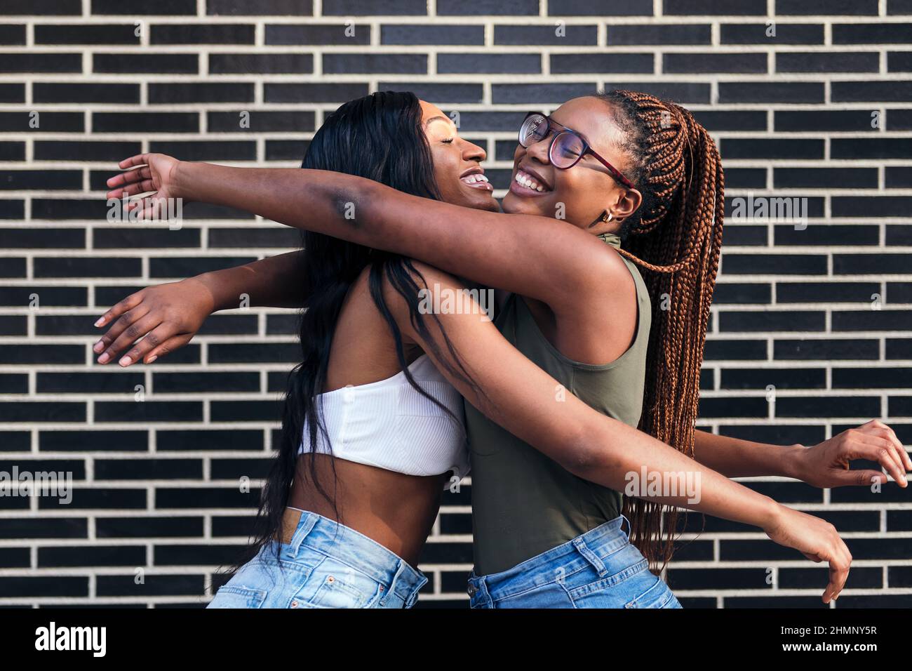 couple of young black girls smiling happy hugging each other in front of a brick wall, concept of youth and friendship, copy space for text Stock Photo