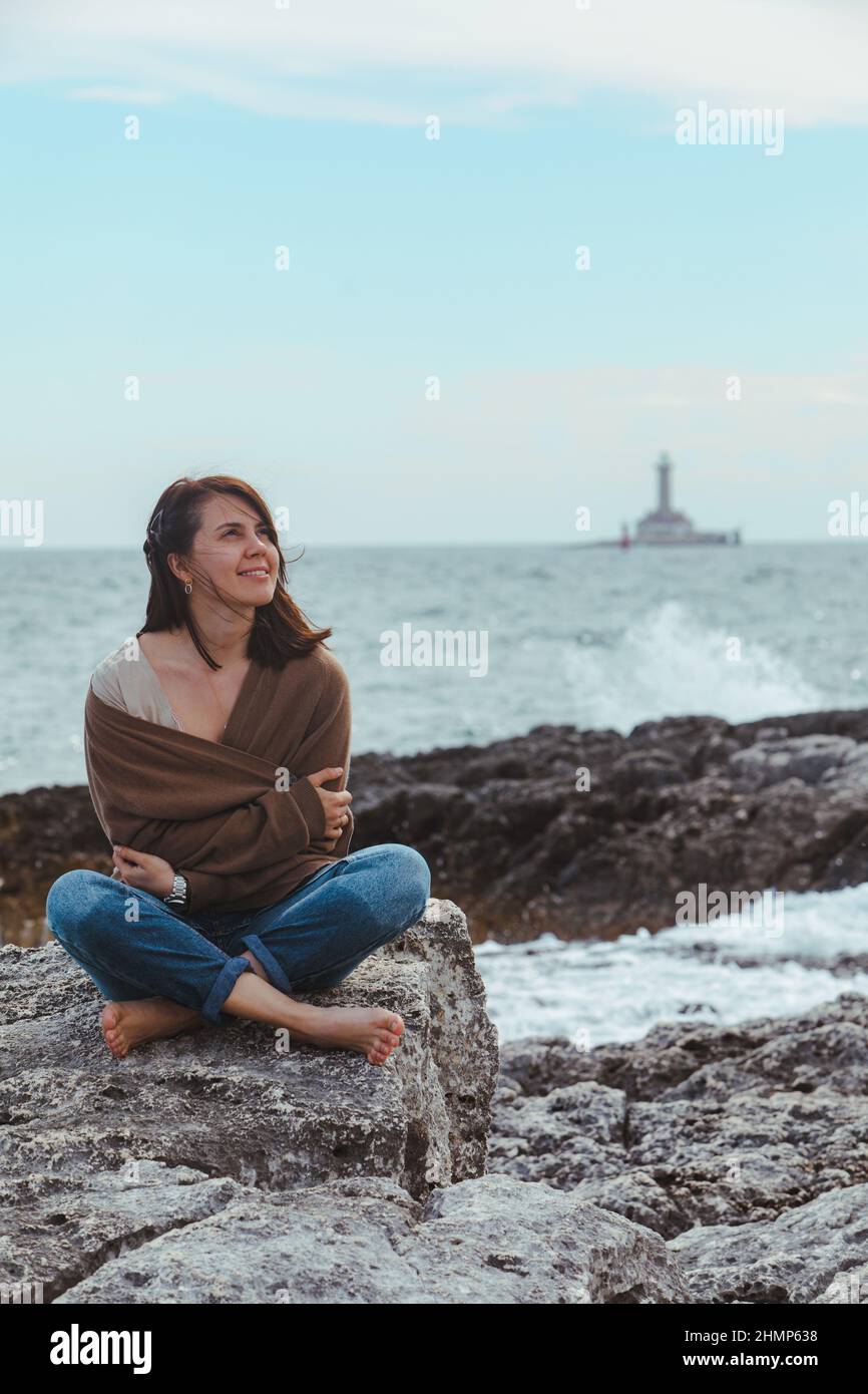 woman sitting by rocky sea beach in wet jeans lighthouse on background.  windy weather. summer vacation. carefree concept Stock Photo - Alamy
