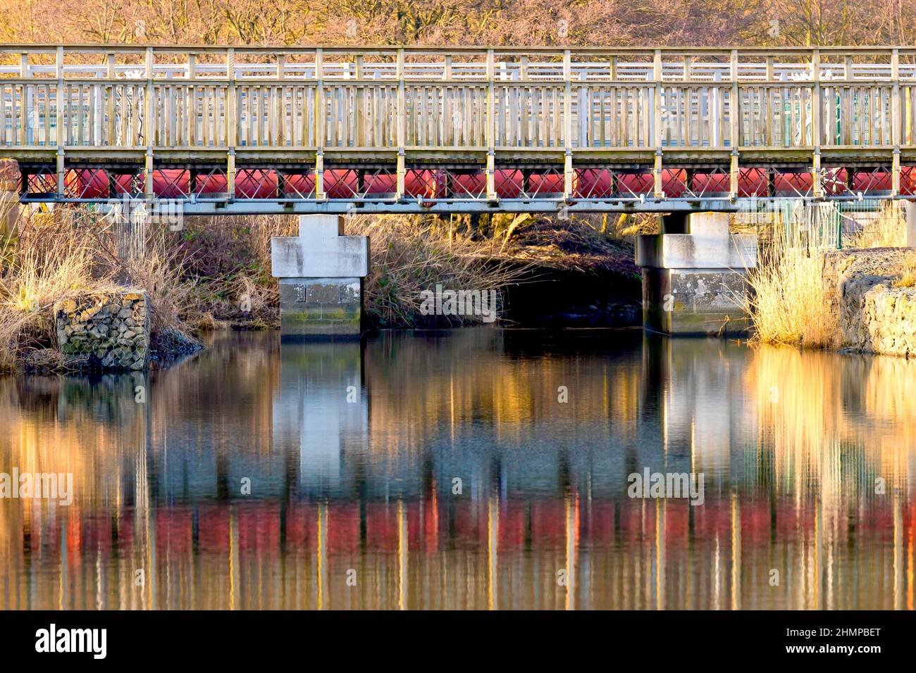 A metal and wood constructed pedestrian foot bridge crossing the Elliot Water, reflected in the calm waters and lit by a low winters afternoon sun. Stock Photo