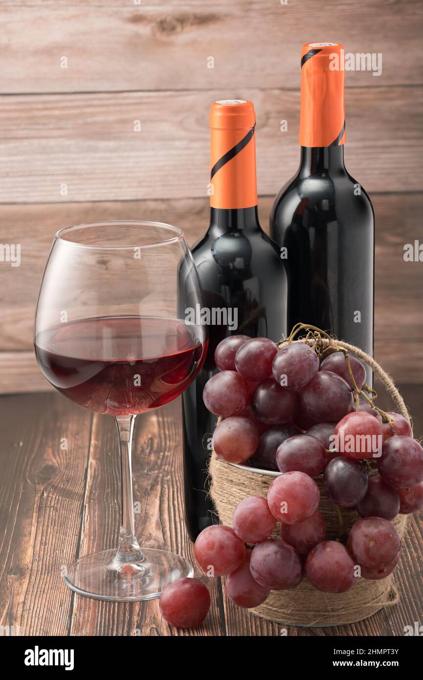 The photo shows two black wine bottles with orange caps. A glass of red wine half filled. Basket with large pink grapes. Composition woody background. Stock Photo