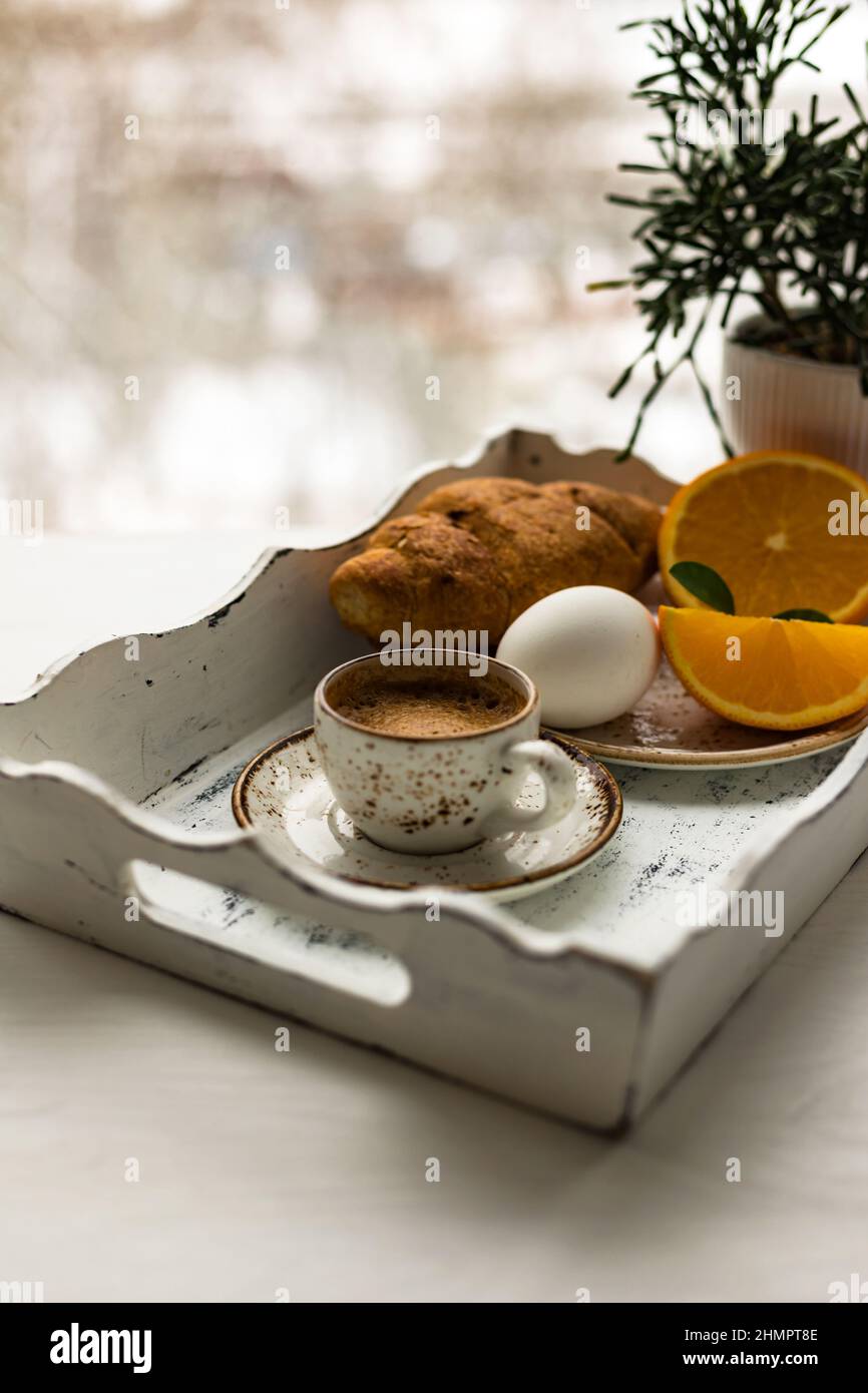 Coffee cup, croissant, egg, fresh orange fruit on white wooden tray against blur window background Stock Photo