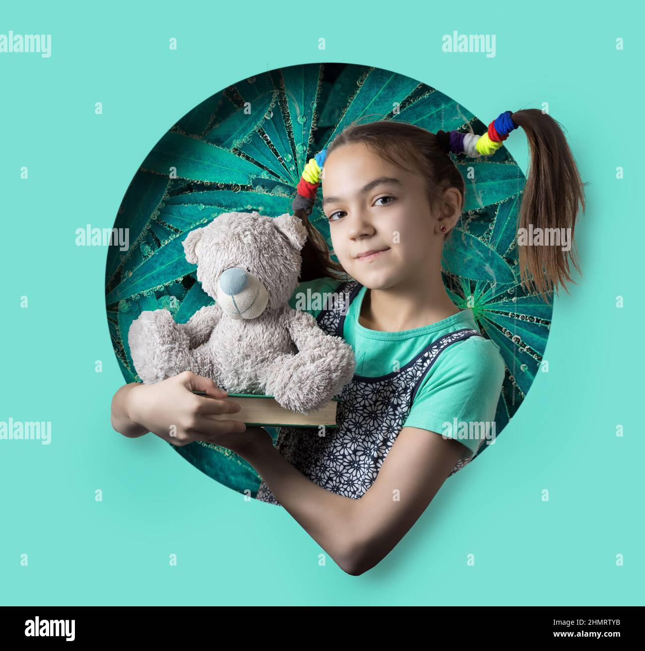 Happy 11 year old girl with funny ponytails holds a book and teddy bear and peeks out of a round hole in paper Stock Photo