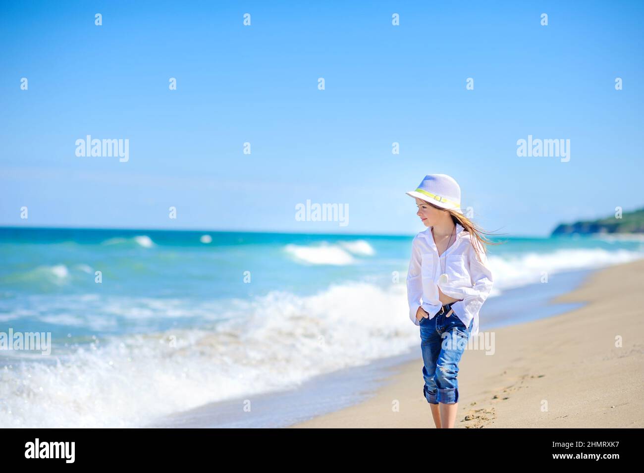 little girl in a beach hat walks along the seashore on a sunny day, healthy rest by the sea Stock Photo