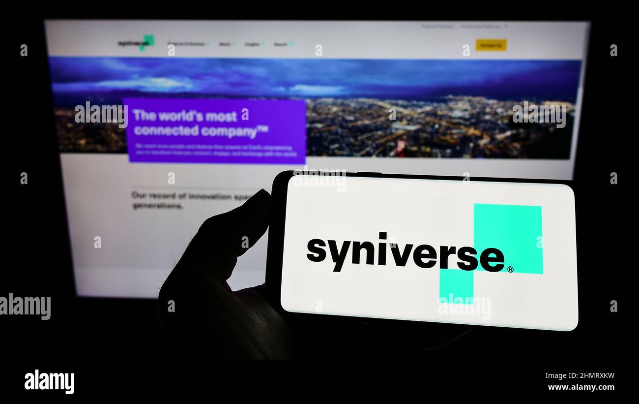 Person holding mobile phone with logo of US telecom company Syniverse Technologies LLC on screen in front of web page. Focus on phone display. Stock Photo