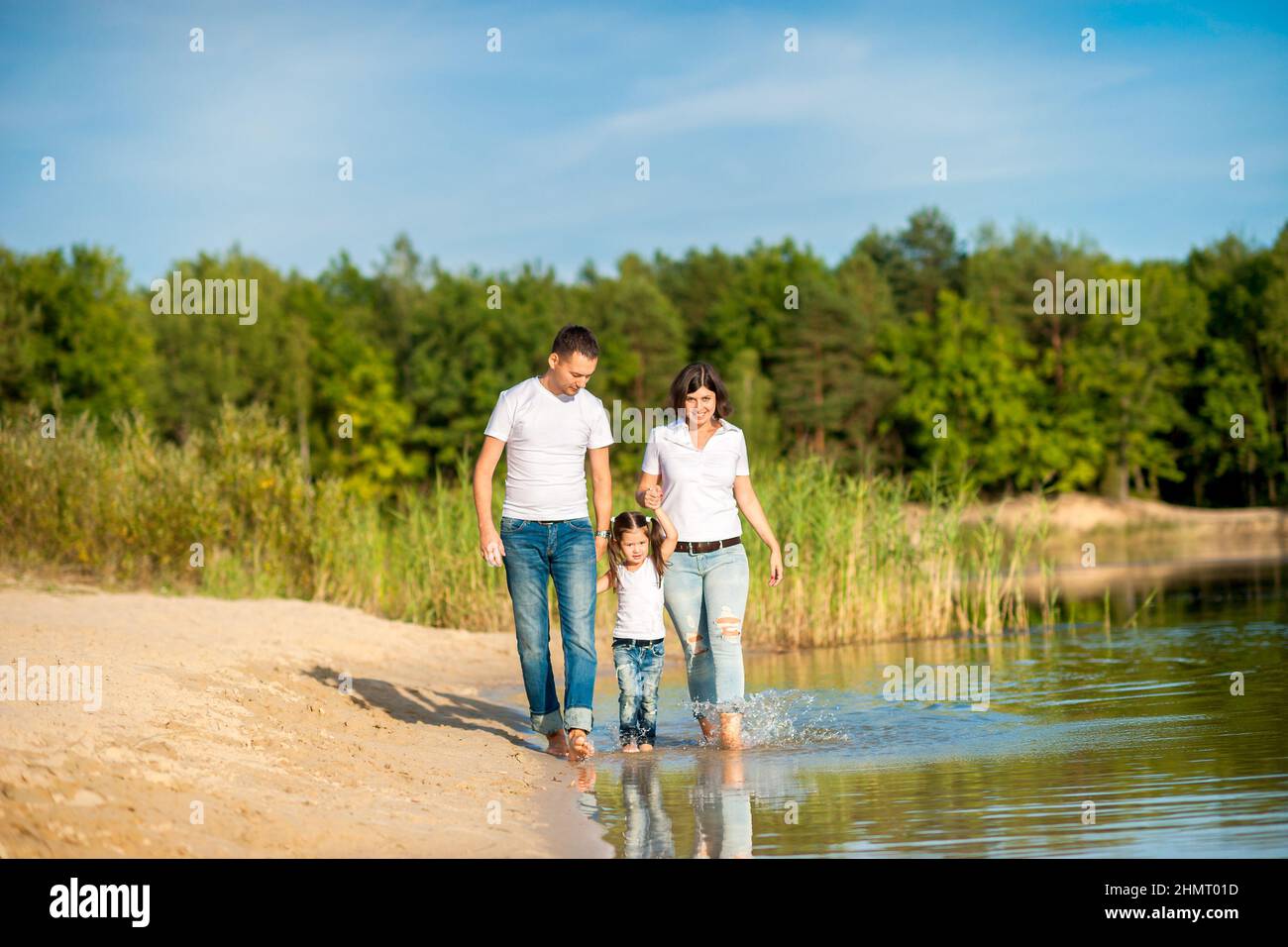 A happy family walks along the river bank on a sunny day, will barefoot on the water and splash, healthy outdoor recreation Stock Photo