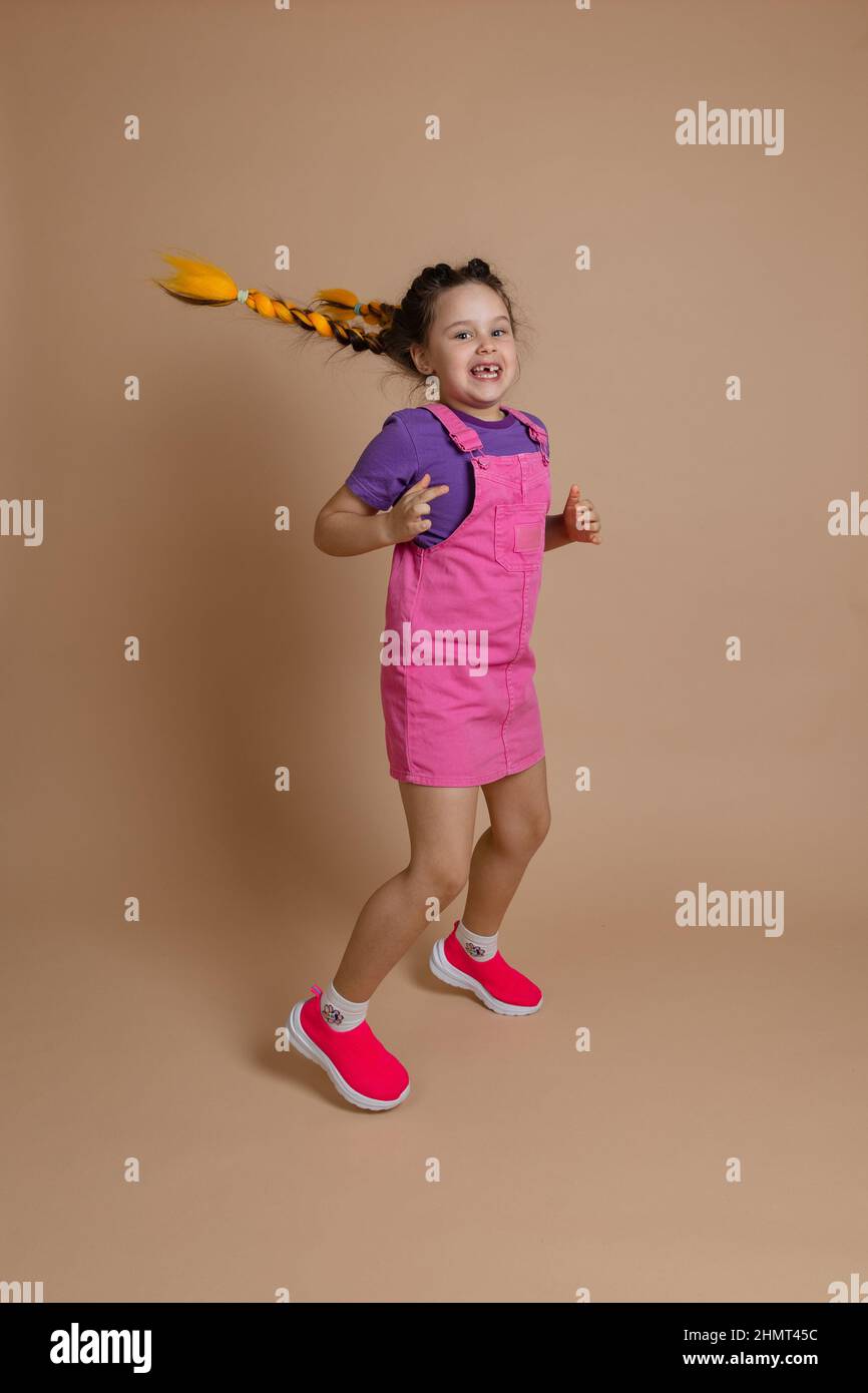 Exuberant active jumping small female kid with two yellow kanekalon pigtails looking at camera with smile with missing tooth in pink jumpsuit, purple Stock Photo