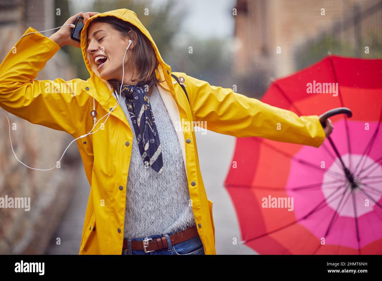 Close-up of a young cheerful woman in a yellow raincoat who is enjoying while listening to the music and walking the city in a good mood on a rainy da Stock Photo