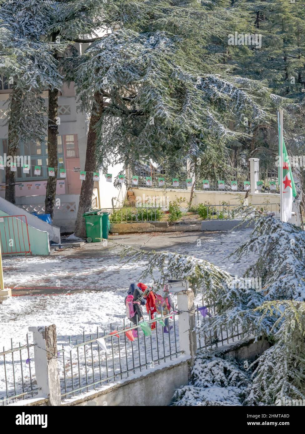 Unrecognizable little girls wearing hood coats carrying their backpacks and waiting outside their school. Ground and trees full of white snow. Stock Photo