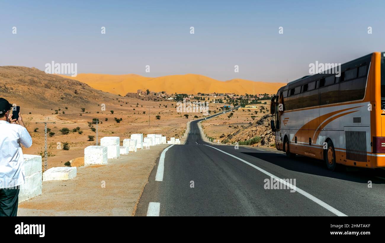 A bus parked and a tourist taking a photo with smartphone of the entrance to Taghit sand dune and reg mountain, white concrete studs along the road. Stock Photo