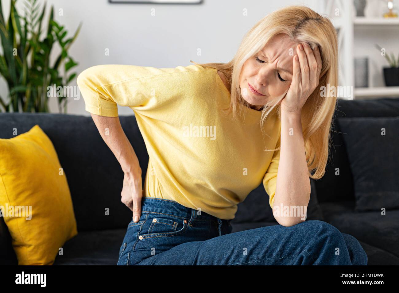 Blonde-haired woman felt a sharp pain in loin sitting on the sofa at home, worried caucasian lady holding a hand on the lower back with suffering face expression Stock Photo