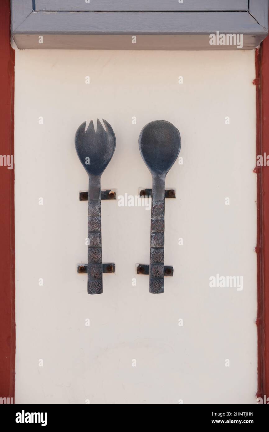 Large metal and old spoon and fork with carvings and patterns on them hang on the wall of cafe or restaurant on Santorini island, Oia, Greece. Signboa Stock Photo