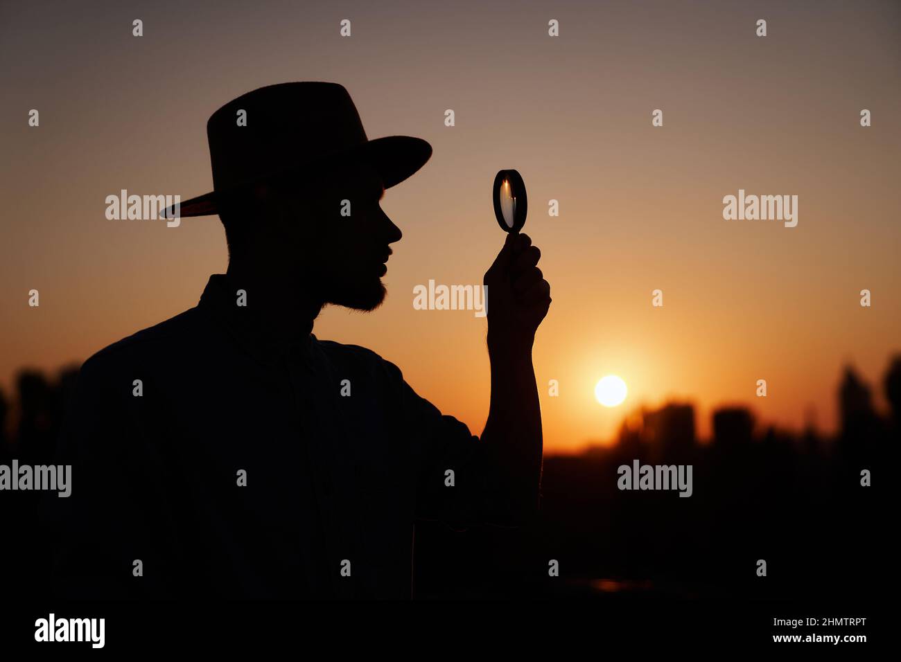 Silhouette of man in hat looking with magnifying at side. Man searching sales or discounts at sunset using loupe with sun on background and urban view. Investigation concept. High quality image Stock Photo