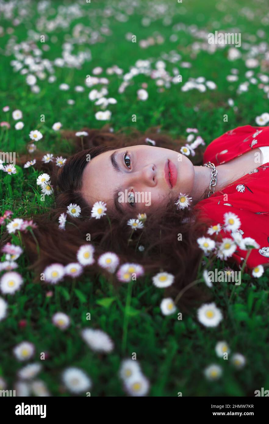 Young beautiful girl in red dress is resting on fresh spring grass with little white flowers. Stock Photo