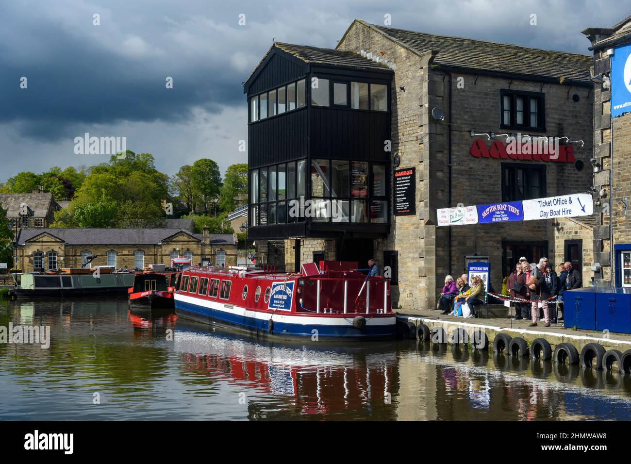 Men women queue for tourist leisure experience on red narrow boat (ticket office, canalside moorings) - Leeds-Liverpool Canal, Yorkshire, England UK. Stock Photo