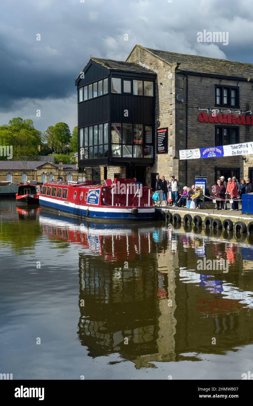 Men women queue for tourist leisure experience on red narrow boat (ticket office, canalside moorings) - Leeds-Liverpool Canal, Yorkshire, England UK. Stock Photo