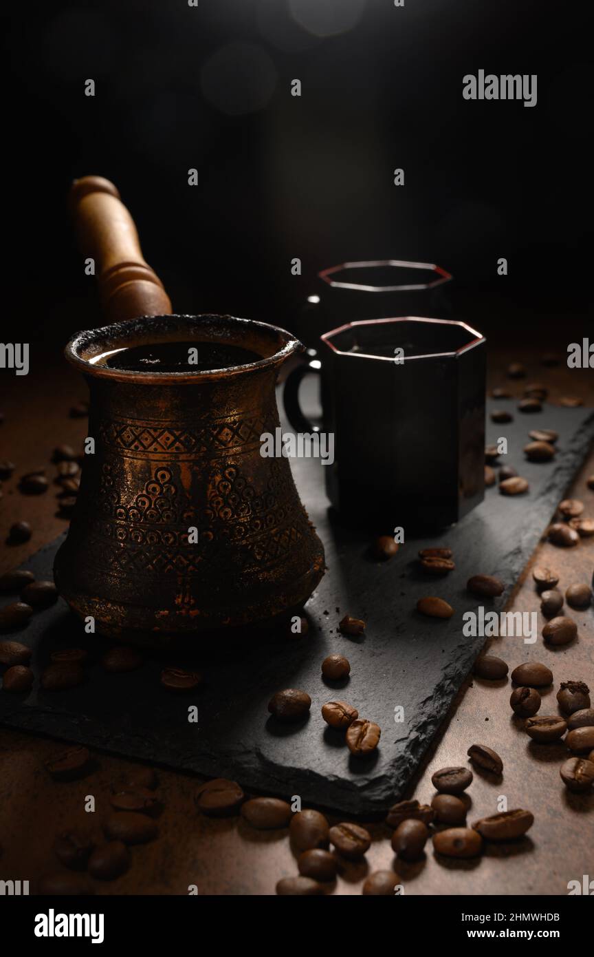 Hot coffee in a Turk. Morning coffee is served on a black board. On the right are two cups of the drink. Black coffee beans are scattered all over the Stock Photo