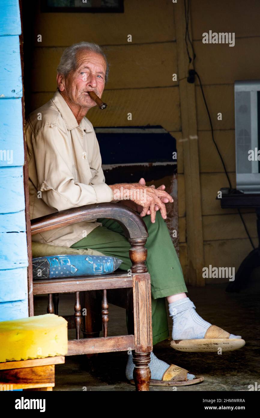 Older man smoking a cuban cigar while sitting on a chair on the front porch of his house in Trinidad, Cuba a Unesco Heritage site. Stock Photo