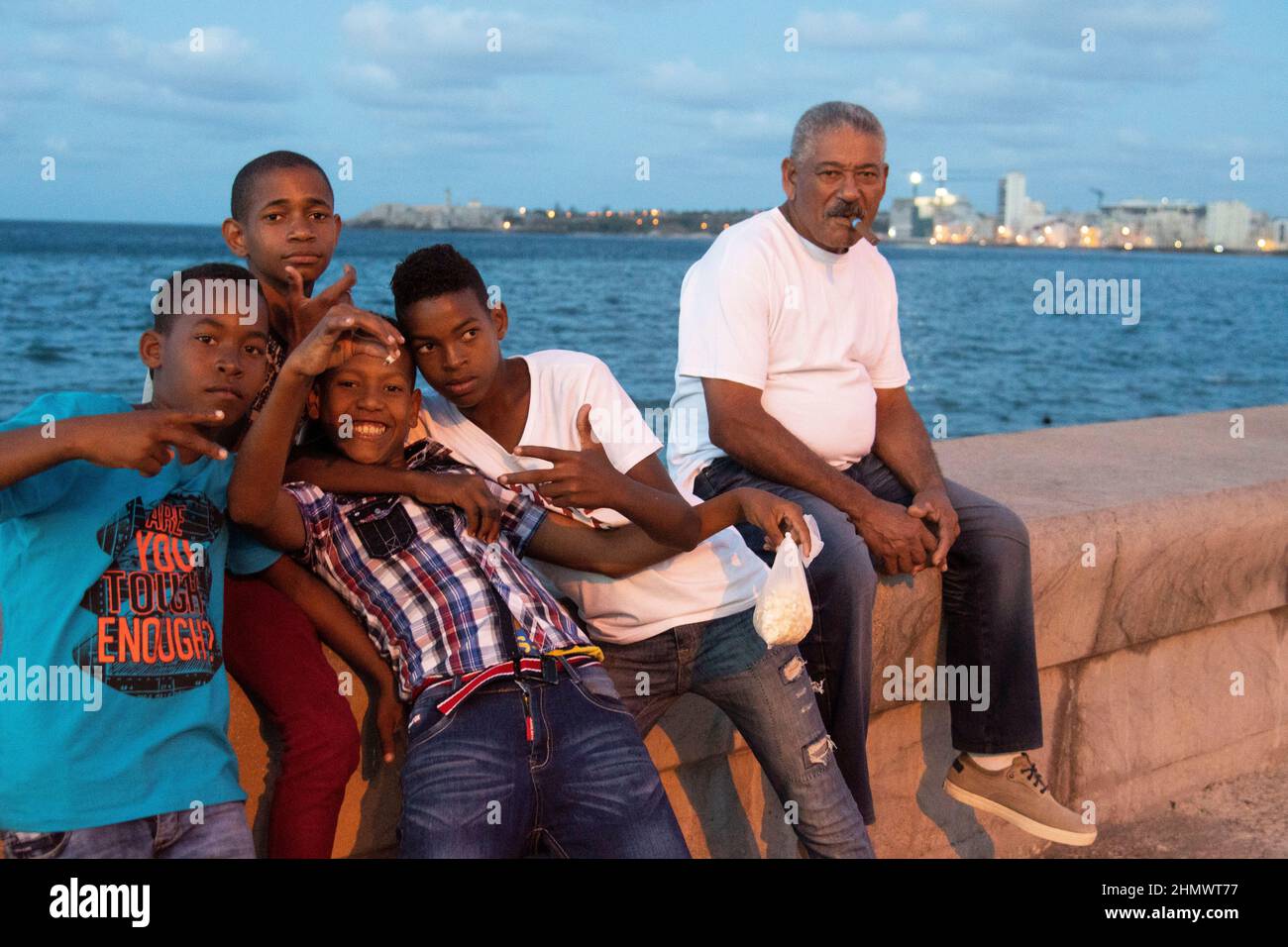 Man smoking cigar sitting with young boy, one holding a bag of popcorn on the malecon seafront in Havana, Cuba. Stock Photo