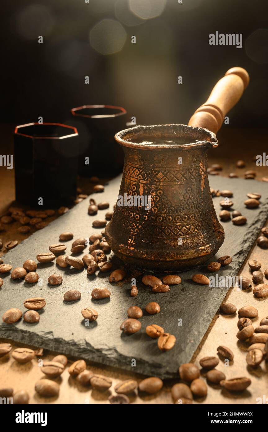 Hot coffee in a Turk. Morning coffee is served on a black board. On the right are two cups of the drink. Black coffee beans are scattered all over the Stock Photo