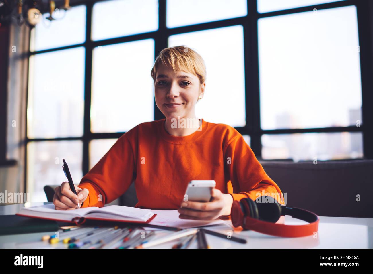 Happy female student writing in notepad and using smartphone in workspace Stock Photo