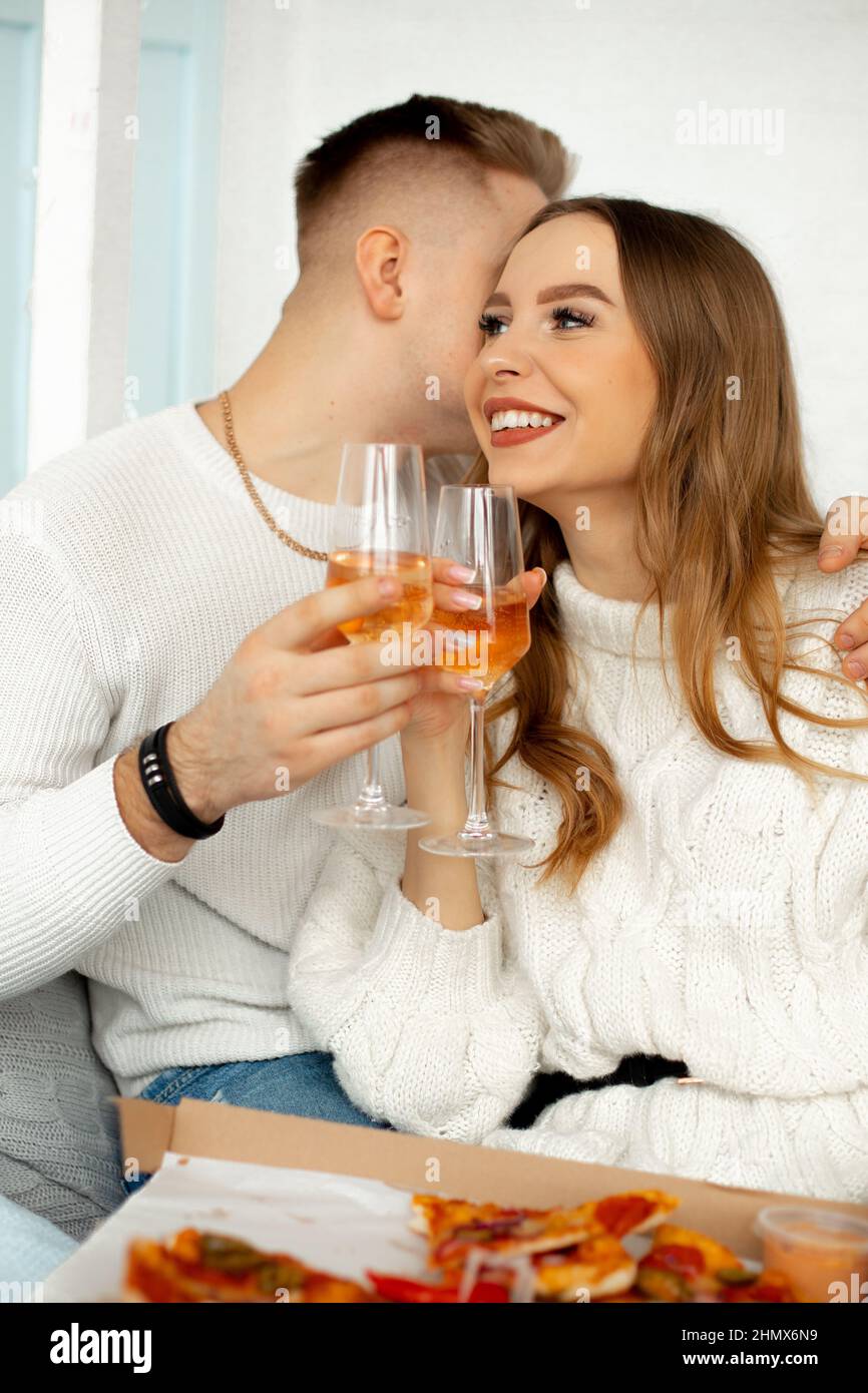 Two tall glass glasses with wine in hands of joyful couple in love. Young people in white knitted sweaters hug, man whispers something tender in ear Stock Photo