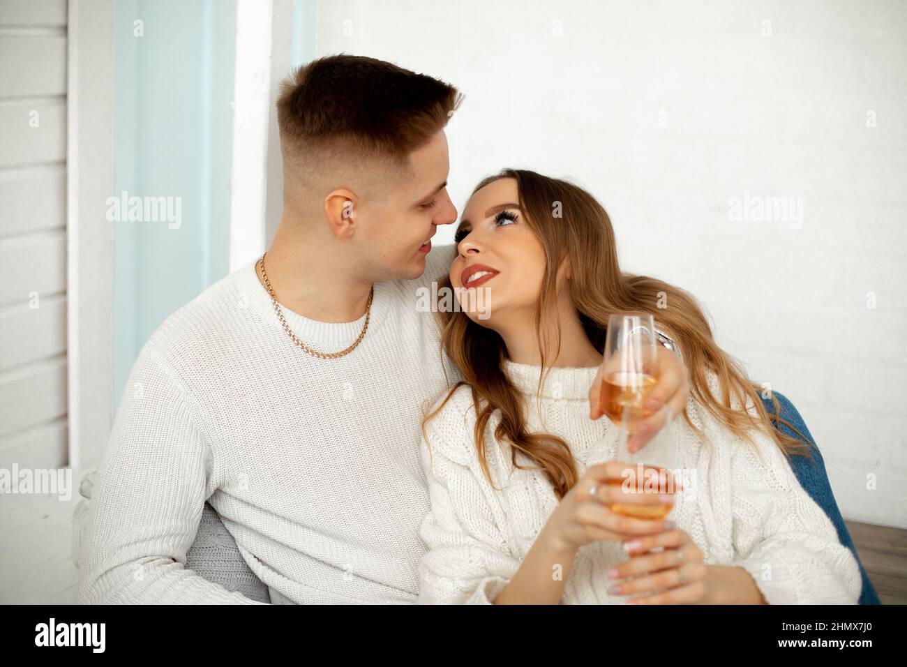 Young girl with brown hair looks at her lover, pressing her back to him, he hugs her shoulders. They are sitting in room with glasses of wine Stock Photo