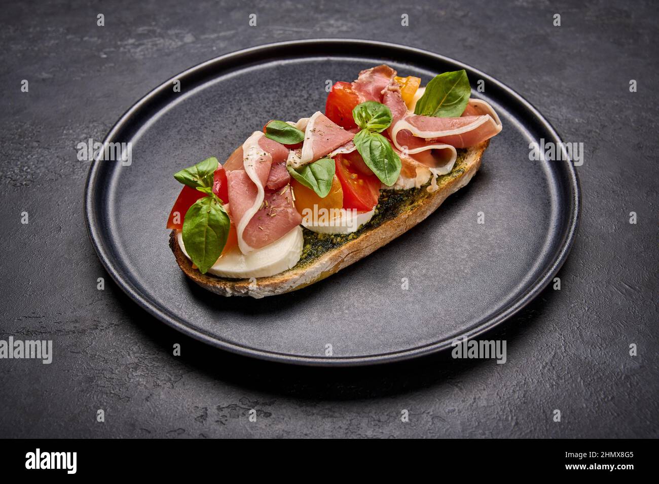 Close up bruschetta with mozzarella, Parma ham and pesto on black plate on graphite background Stock Photo