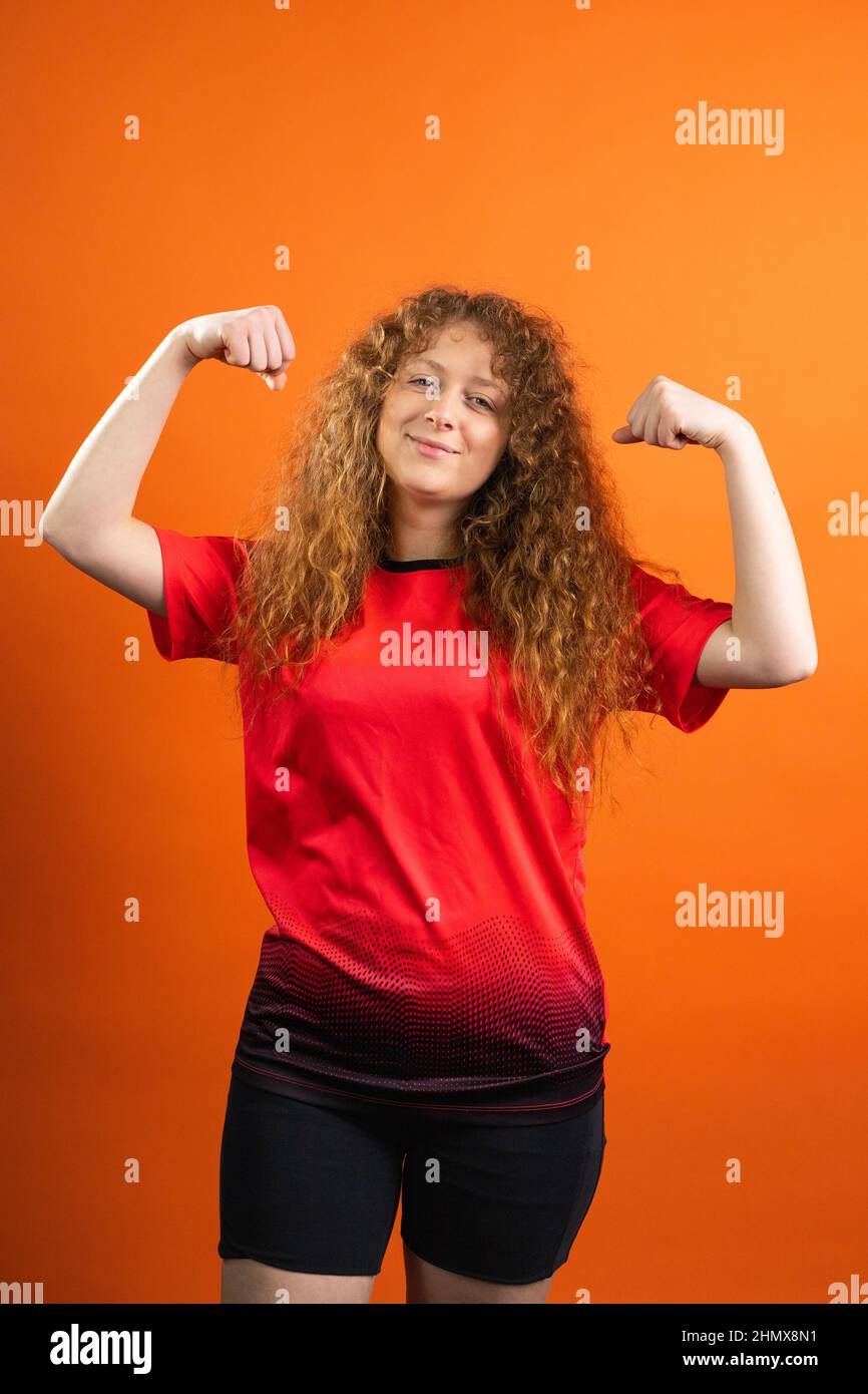 Strong redhead young woman fan of football in team uniform red and black color posing for the camera with satisfied facial expression. Stock Photo