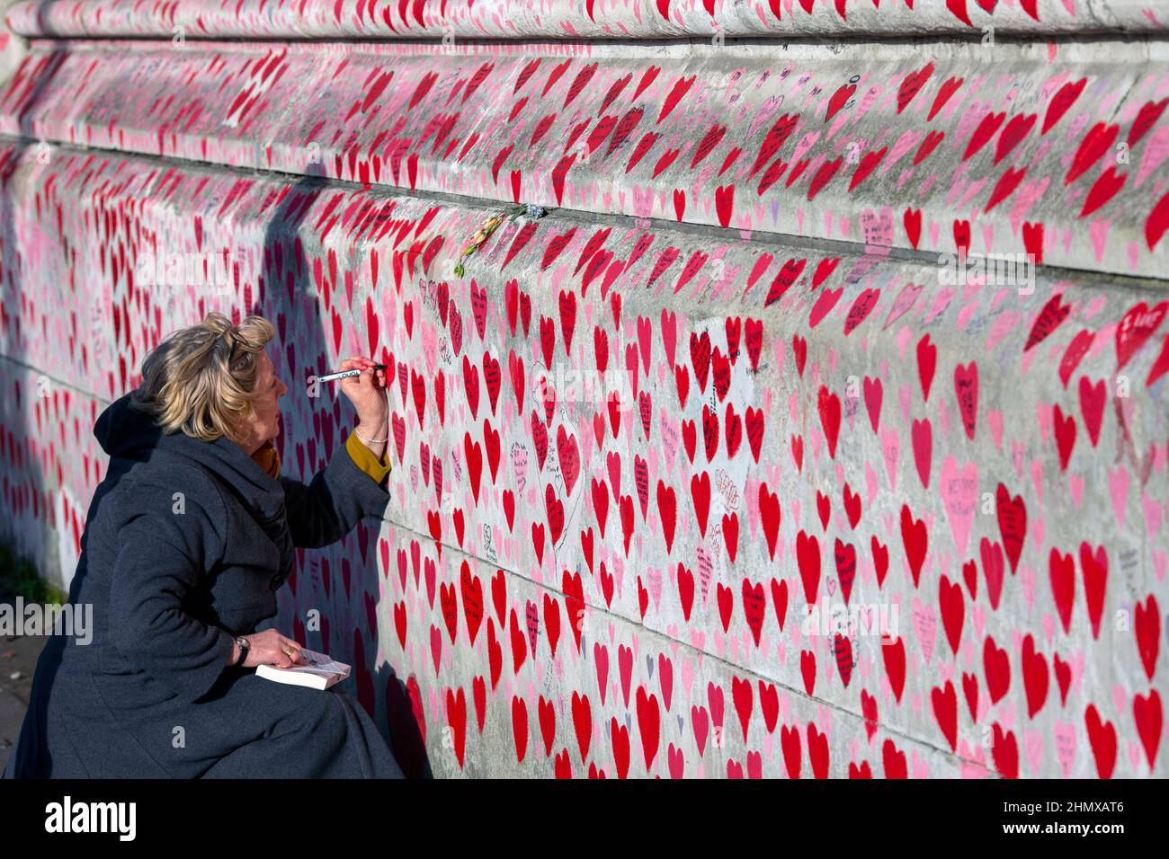 Female writing names on The National COVID Memorial Wall painted with red hearts on beautiful sunny day . Westminster, London , UK . Stock Photo