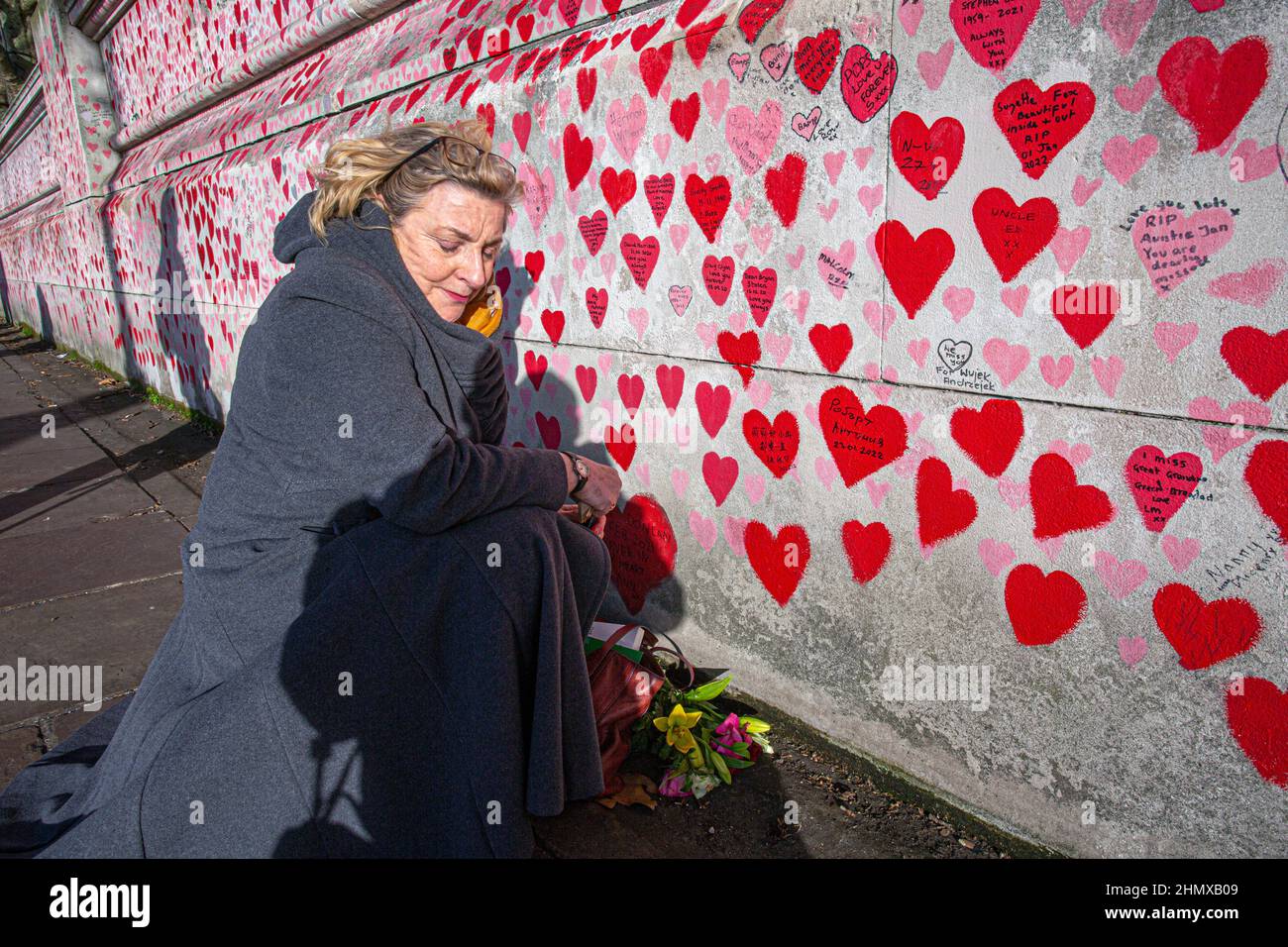 Female writing names on The National COVID Memorial Wall painted with red hearts on beautiful sunny day . Westminster, London , UK . Stock Photo