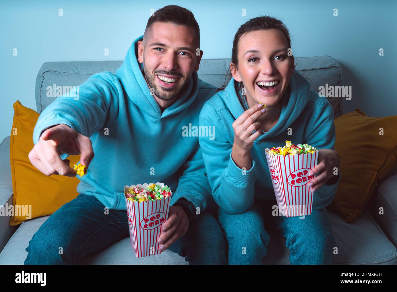 Young happy couple with popcorn sitting on sofa and watching funny comedy movie. Couple enjoying time together at home Stock Photo