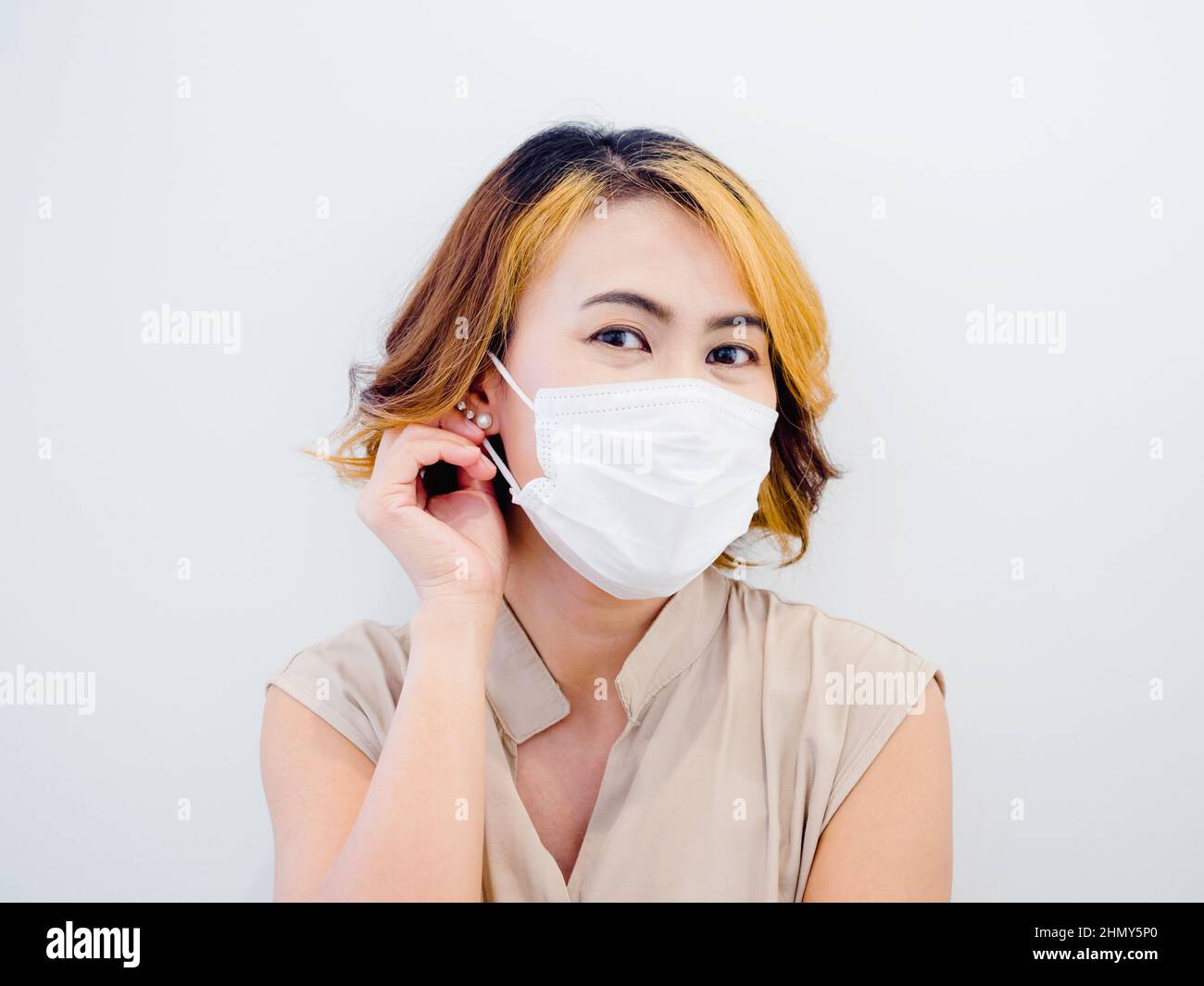 Beauty Asian woman in a beige sleeveless shirt with short brown hair and highlights wearing a white protective face mask isolated on a white backgroun Stock Photo