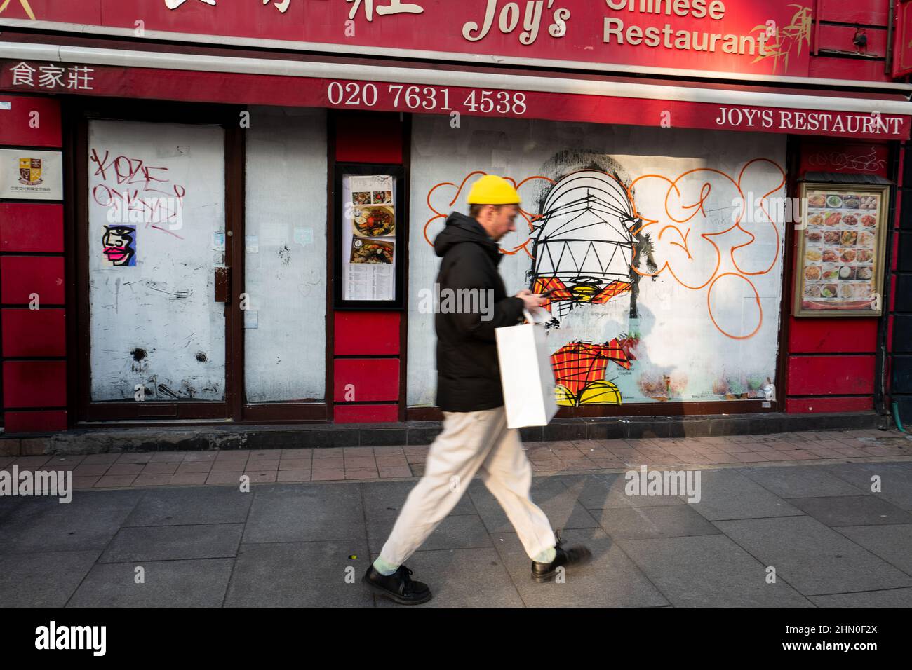Man wearing a yellow at walks past a closed down Chinese Restaurant on New Oxford Street, London UK. Graffiti by Nathan Bowen. Stock Photo