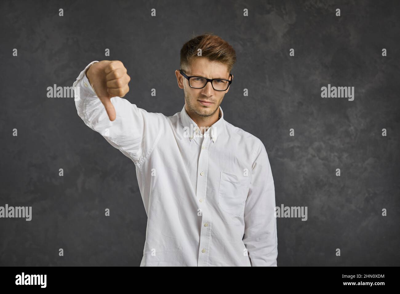 Dissatisfied and frustrated young man showing thumb down standing on gray background. Stock Photo