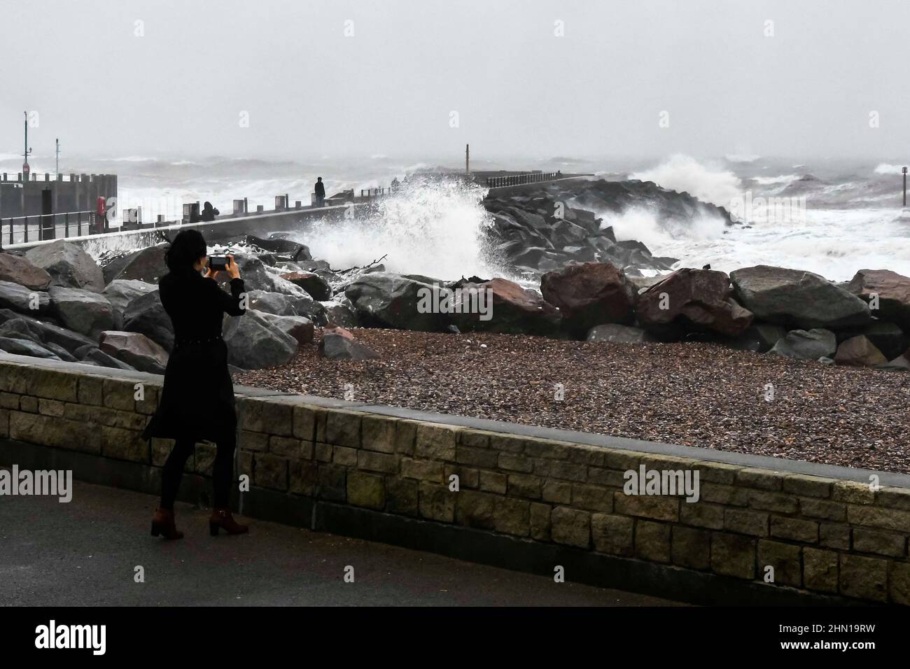 West Bay Dorset, UK.  13th February 2022.  UK Weather.  A woman photographs the stormy sea crash against the sea defences on her phone at West Bay in Dorset on an afternoon of heavy rain and gale force winds.  Picture Credit: Graham Hunt/Alamy Live News Stock Photo