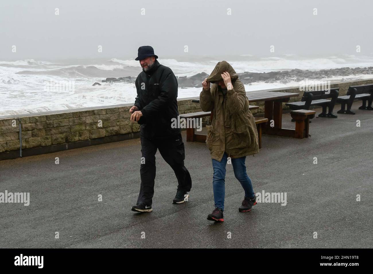 West Bay Dorset, UK.  13th February 2022.  UK Weather.  A couple struggle to walk in the strong gusty wind as stormy sea crash against the sea defences at West Bay in Dorset on an afternoon of heavy rain and gale force winds.  Picture Credit: Graham Hunt/Alamy Live News Stock Photo