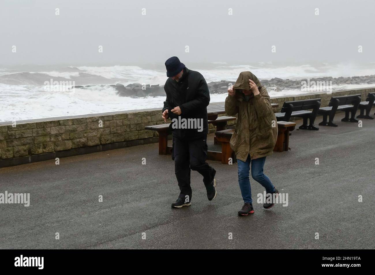 West Bay Dorset, UK.  13th February 2022.  UK Weather.  A couple struggle to walk in the strong gusty wind as stormy sea crash against the sea defences at West Bay in Dorset on an afternoon of heavy rain and gale force winds.  Picture Credit: Graham Hunt/Alamy Live News Stock Photo