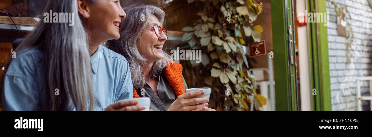 Positive mature women companions with cups sit near window of cafe Stock Photo